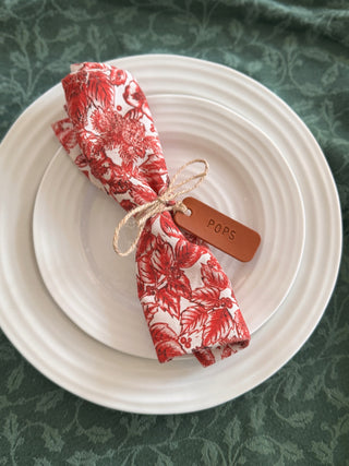 Red floral napkin on a white plate with a tan leather name tag on a green patterned background