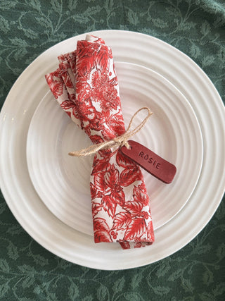 A folded red and white floral patterned napkin tied with a twine string and a small wooden tag engraved with the name 'ROSIE', presented on a stack of white plates over a dark green patterned tablecloth.