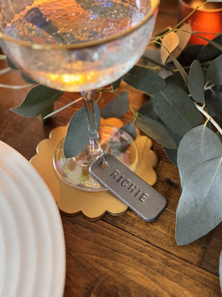 Cocktail glass with a decorative rim on a wooden table, accompanied by a name tag and greenery.