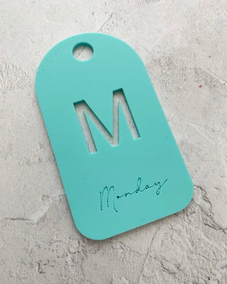 Turquoise tag with letter 'M' and 'Monday' on a gray surface
