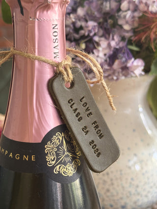 Pink champagne bottle with a 'Love From Class 24 2026' in olive green leather tag against a blurred floral background