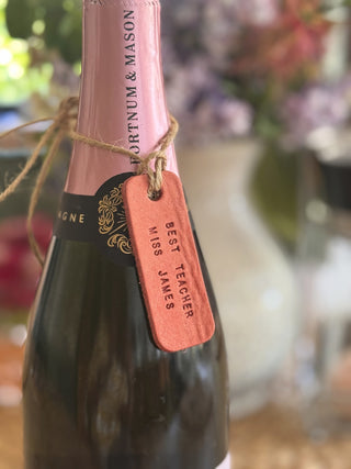 Bottle of Fortnum & Mason champagne with a personalised pink leather tag in a blurred floral setting. Gift for Teacher