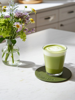 Matcha green tea in a glass on a coaster with a vase of flowers in the background.