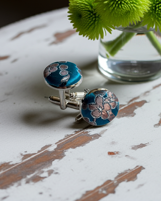 Blue floral cufflinks on a wooden surface with green flowers in the background
