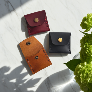 Three small leather pouches in red, navy, and brown on a light surface with a plant in the corner.