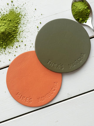 Two round coasters, one green and one orange, with 'Thea's Matcha' and 'Mike's Kombucha' branding on a white surface with matcha powder.