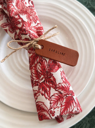 Red floral napkin on a white plate with a name tag, tan leather tag stamped with Caroline
