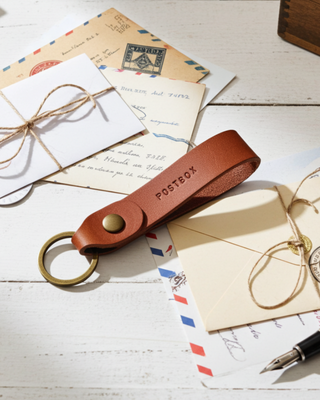 Tan leather keychain with 'POSTBOX' branding on a wooden surface with envelopes and a pen.