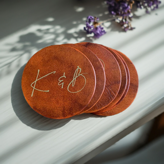 A set of round brown leather coasters with the initials 'K & B' embossed on them, displayed on a white surface with a floral pattern in the background.